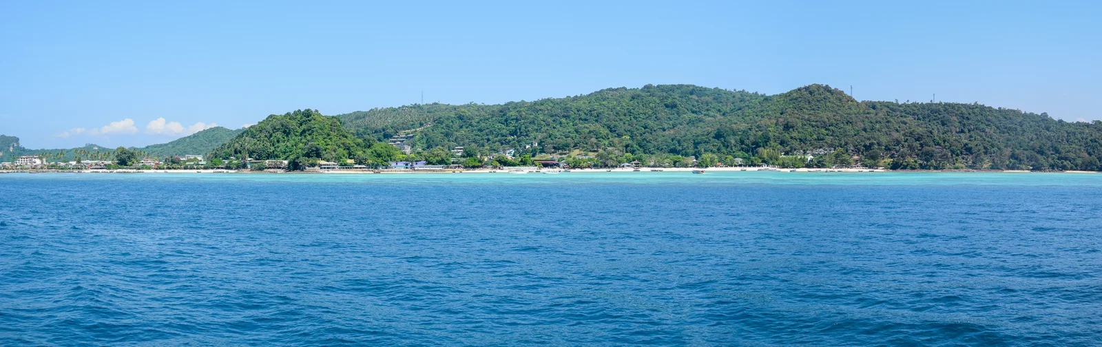 Forested green hills of Ko Phi Phi Don rising behind a low sandy shore lined with buildings, seen across turquoise water from the sea