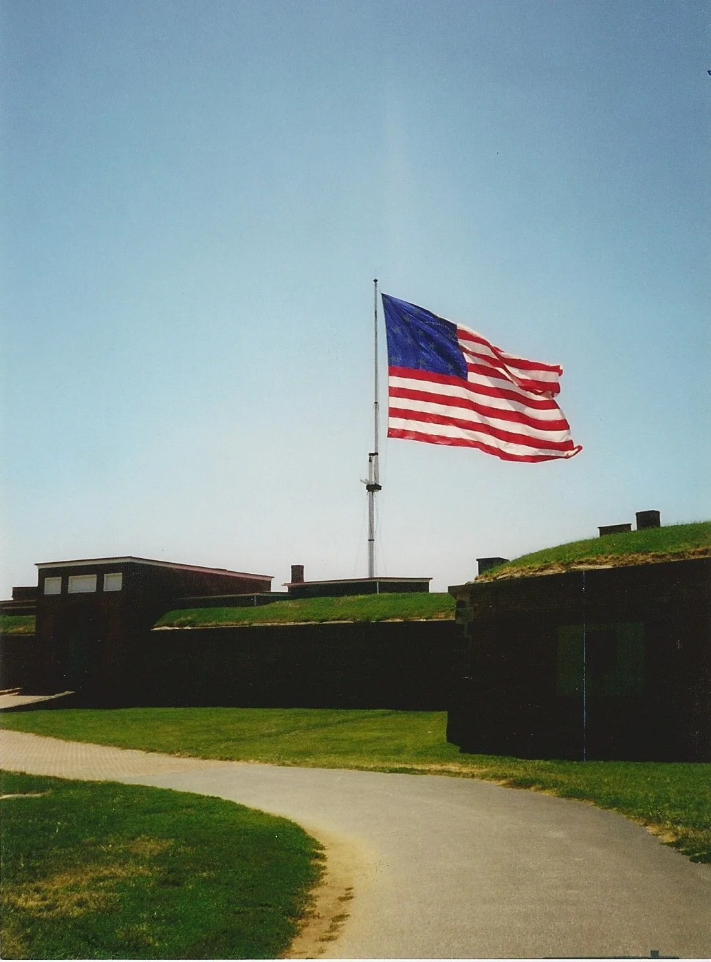 The large fifteen-star, fifteen-stripe garrison flag flying from a tall flagpole above the grass-topped brick ramparts and entrance of Fort McHenry on a clear summer day