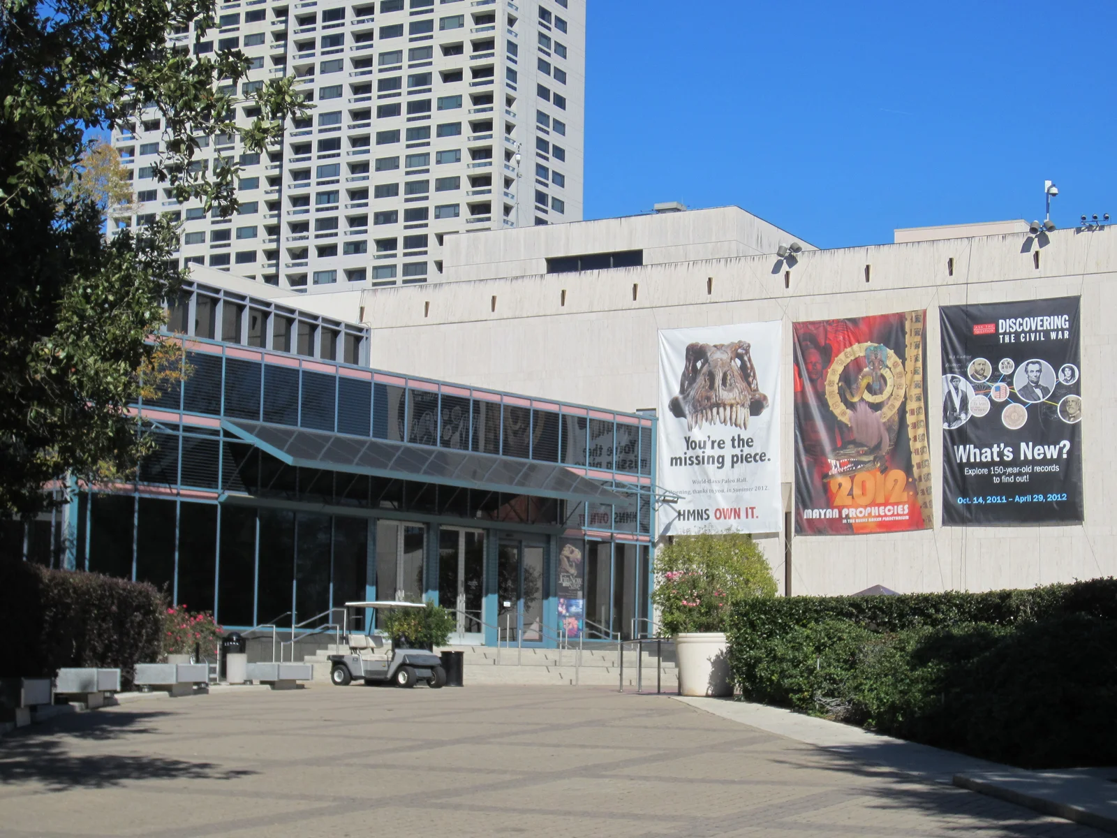 Entrance plaza of the Houston Museum of Natural Science with a glass-walled walkway and large exhibition banners on a sunny day