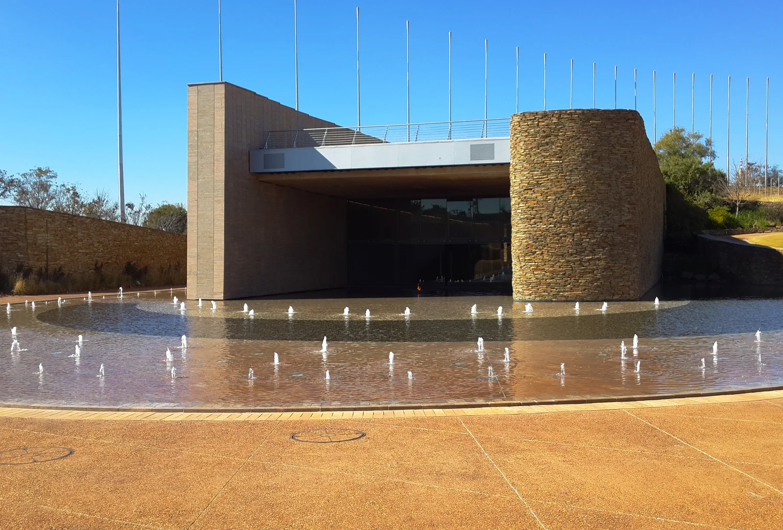 Freedom Park's S'khumbuto stage wall behind a circular pool with rows of fountain jets and tall metal reed poles under a clear blue sky