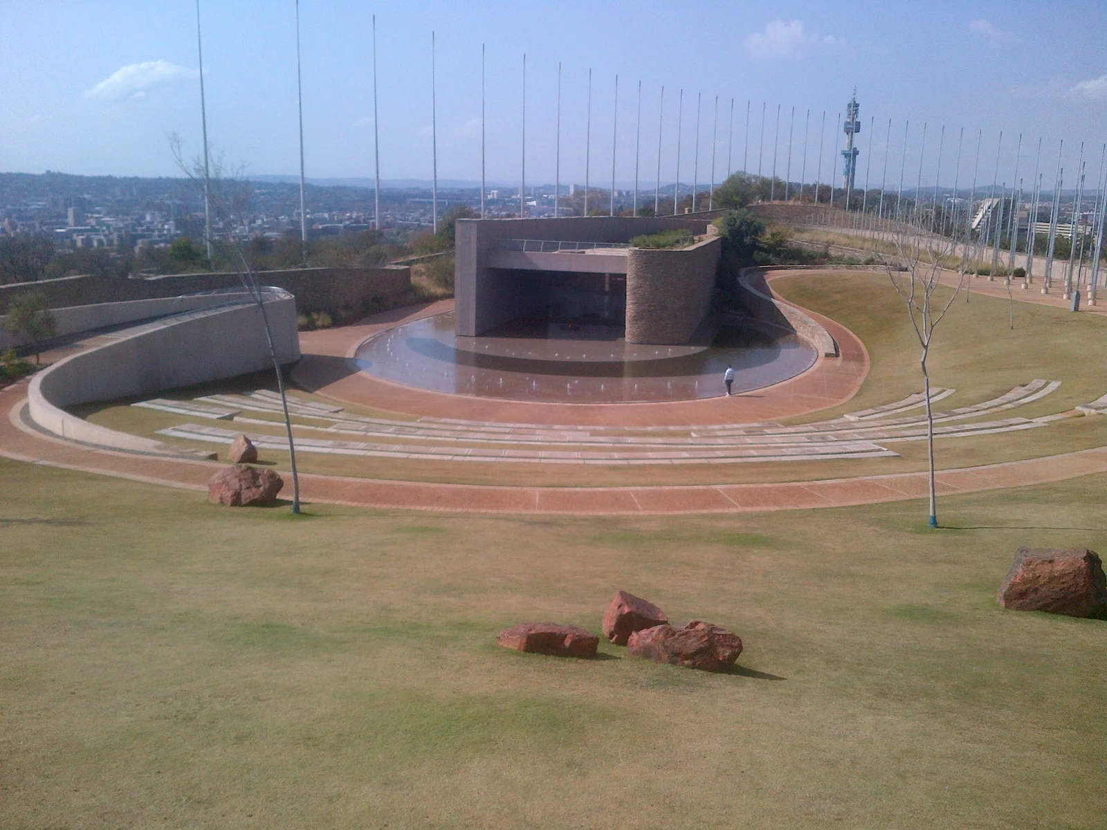 Elevated view over Freedom Park's terraced amphitheatre and reflecting pool, with red boulders scattered on the lawn and the Pretoria skyline on the distant horizon