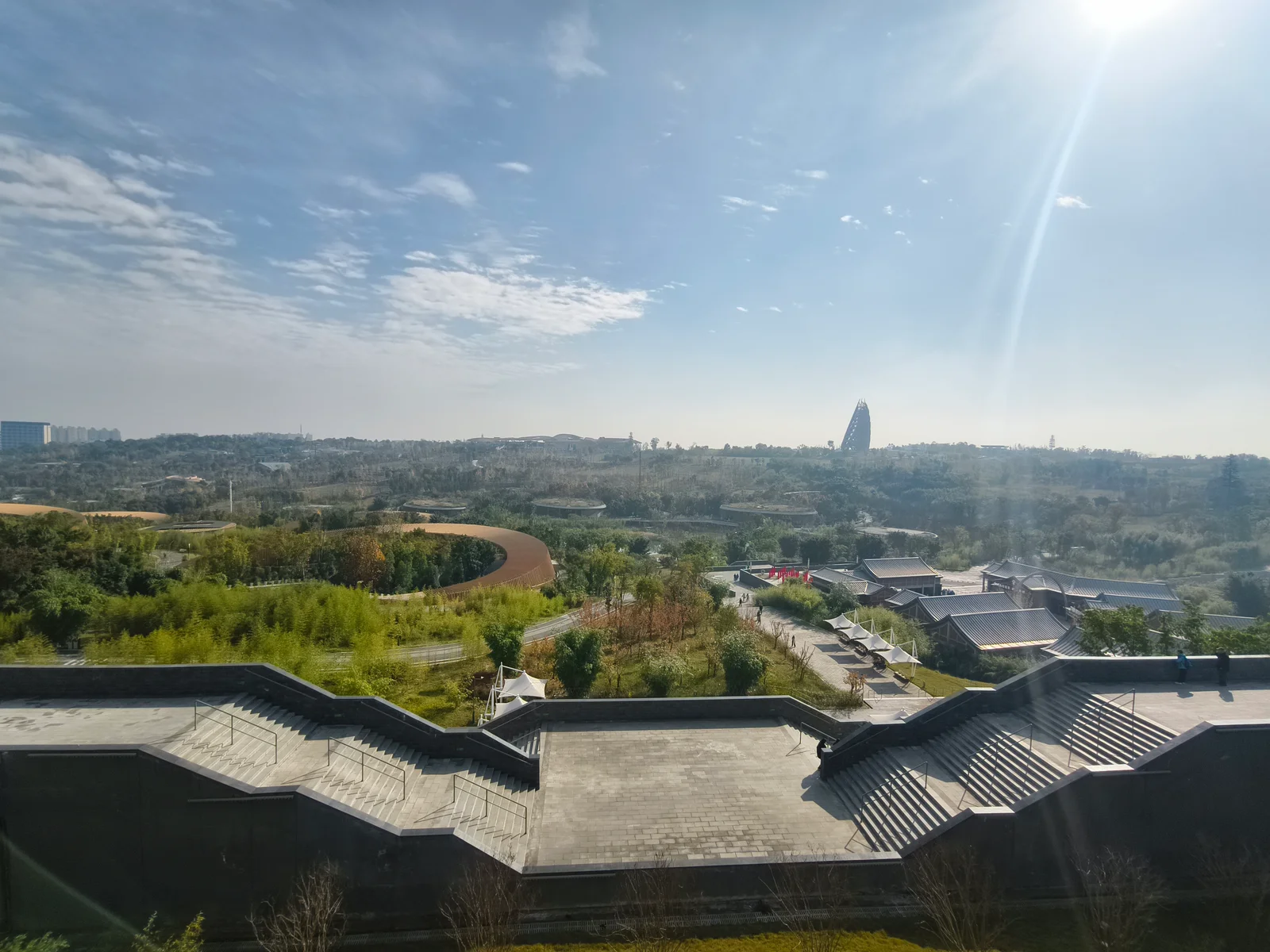 Elevated view over the Chengdu panda base with wide stone staircases, grey-roofed buildings and wooded parkland under a clear blue sky