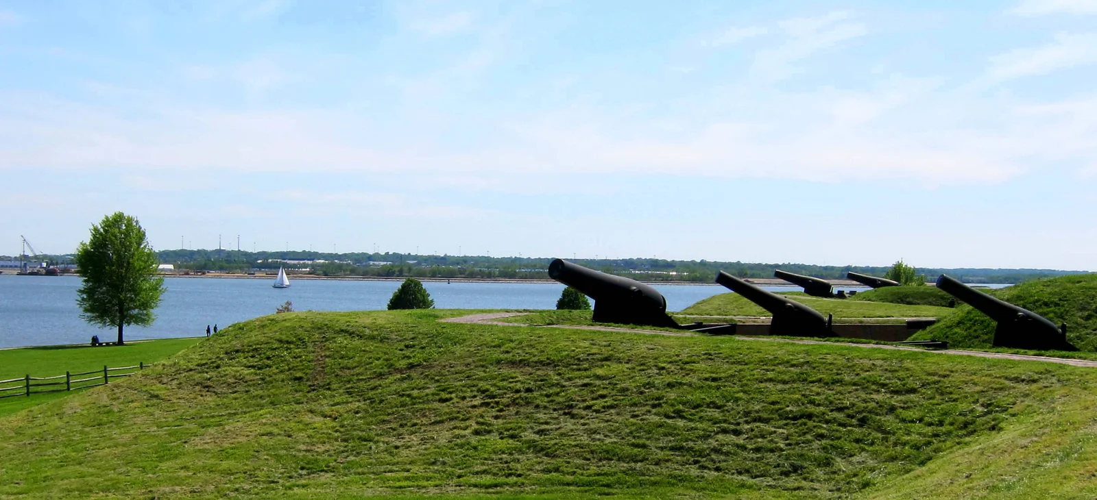 A row of large black iron Rodman cannons mounted along the grassy earthwork rampart of Fort McHenry, their barrels angled out over the Patapsco River with a sailboat on the water beyond