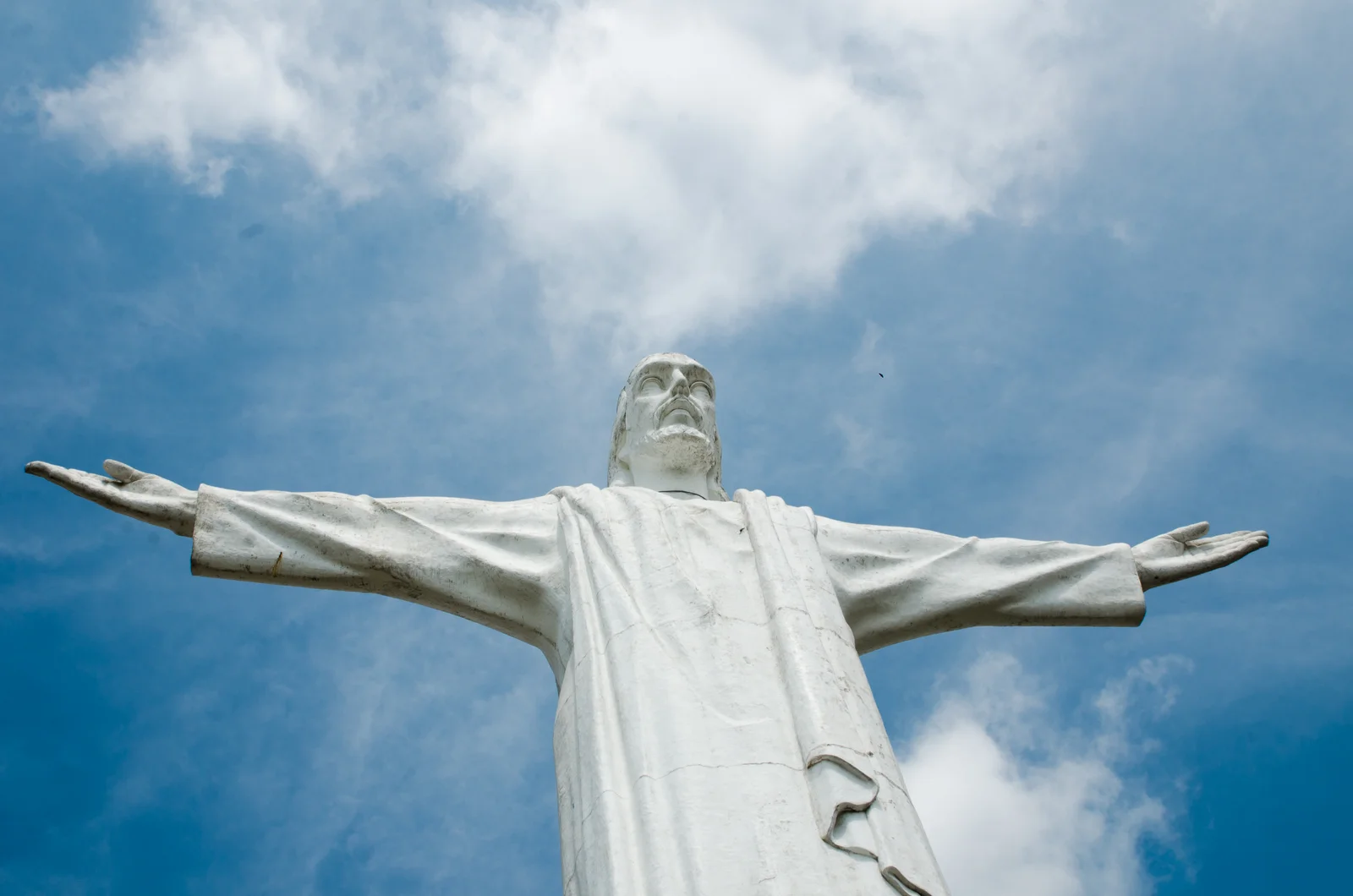 The white Cristo Rey statue seen from directly below, arms outstretched against a blue sky with scattered clouds