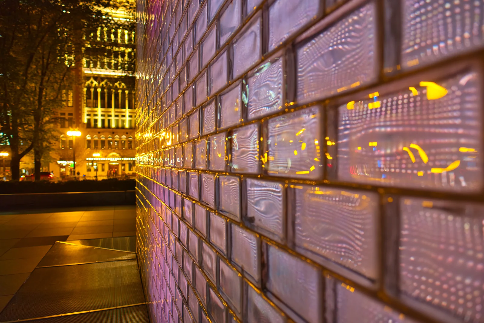 Nighttime close-up of the glass-block face of a Crown Fountain tower glowing pink and gold, receding in perspective toward a lit historic building