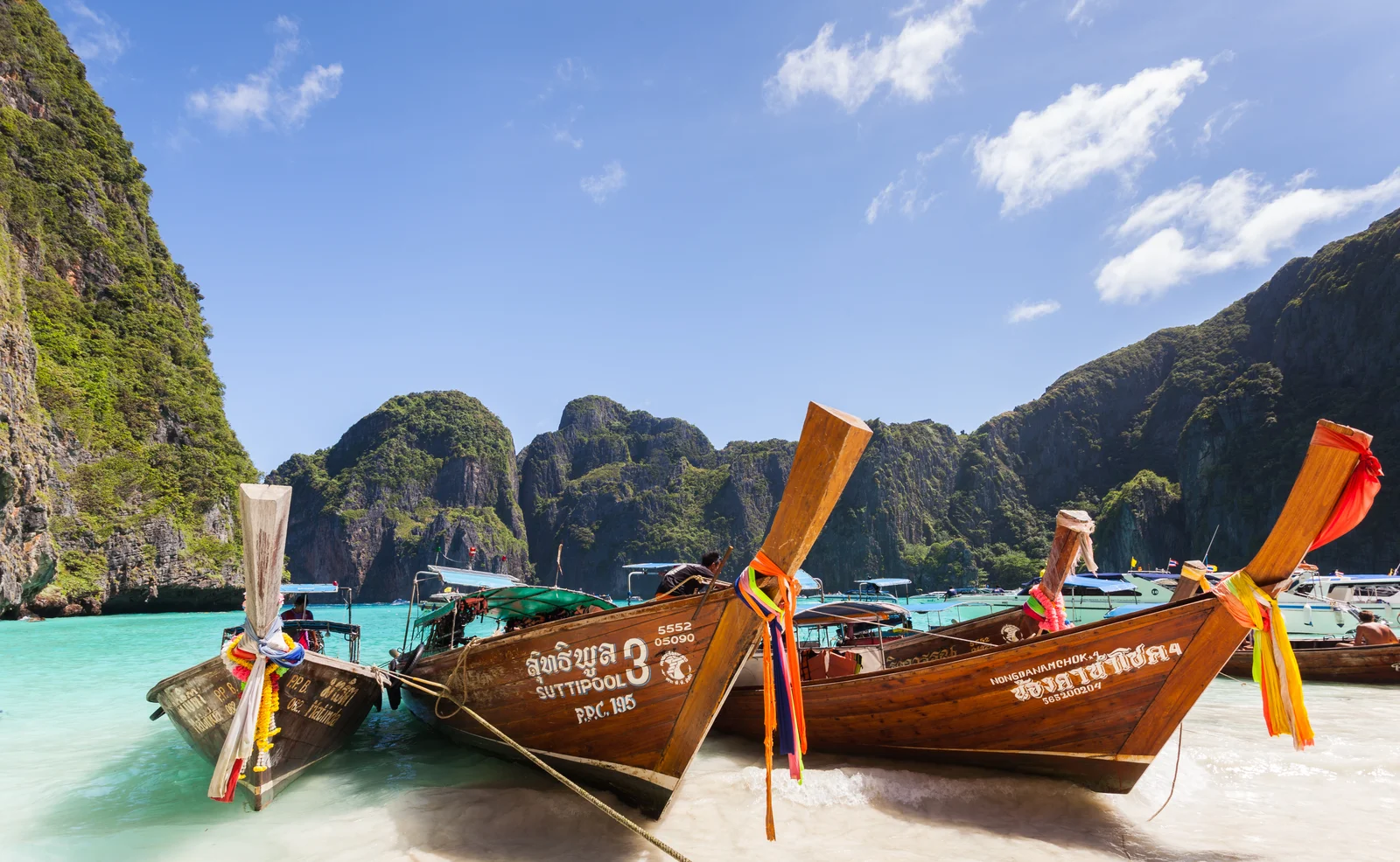 Three wooden long-tail boats moored on the white sand of Maya Bay beneath towering limestone karst cliffs