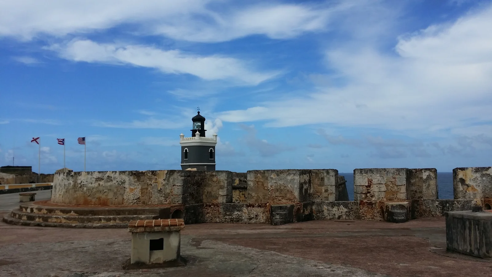 The grey and white El Morro lighthouse tower rising above the fort's weathered upper ramparts, with three flags flying and the Atlantic on the right under a blue sky