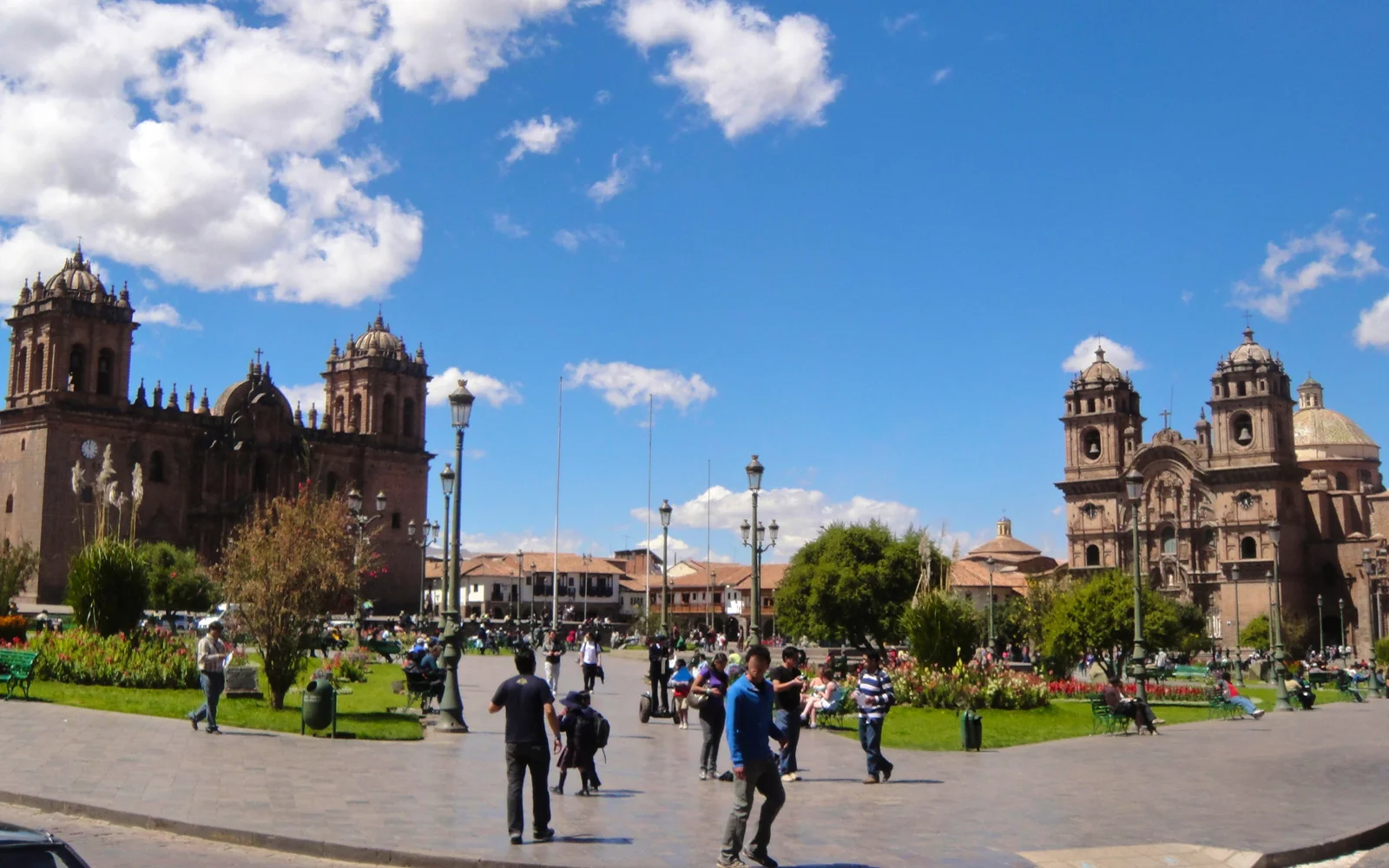 The main square of Cusco with a stone cathedral and its bell towers on the left and a twin-towered baroque church on the right, people crossing the paved plaza under a blue sky