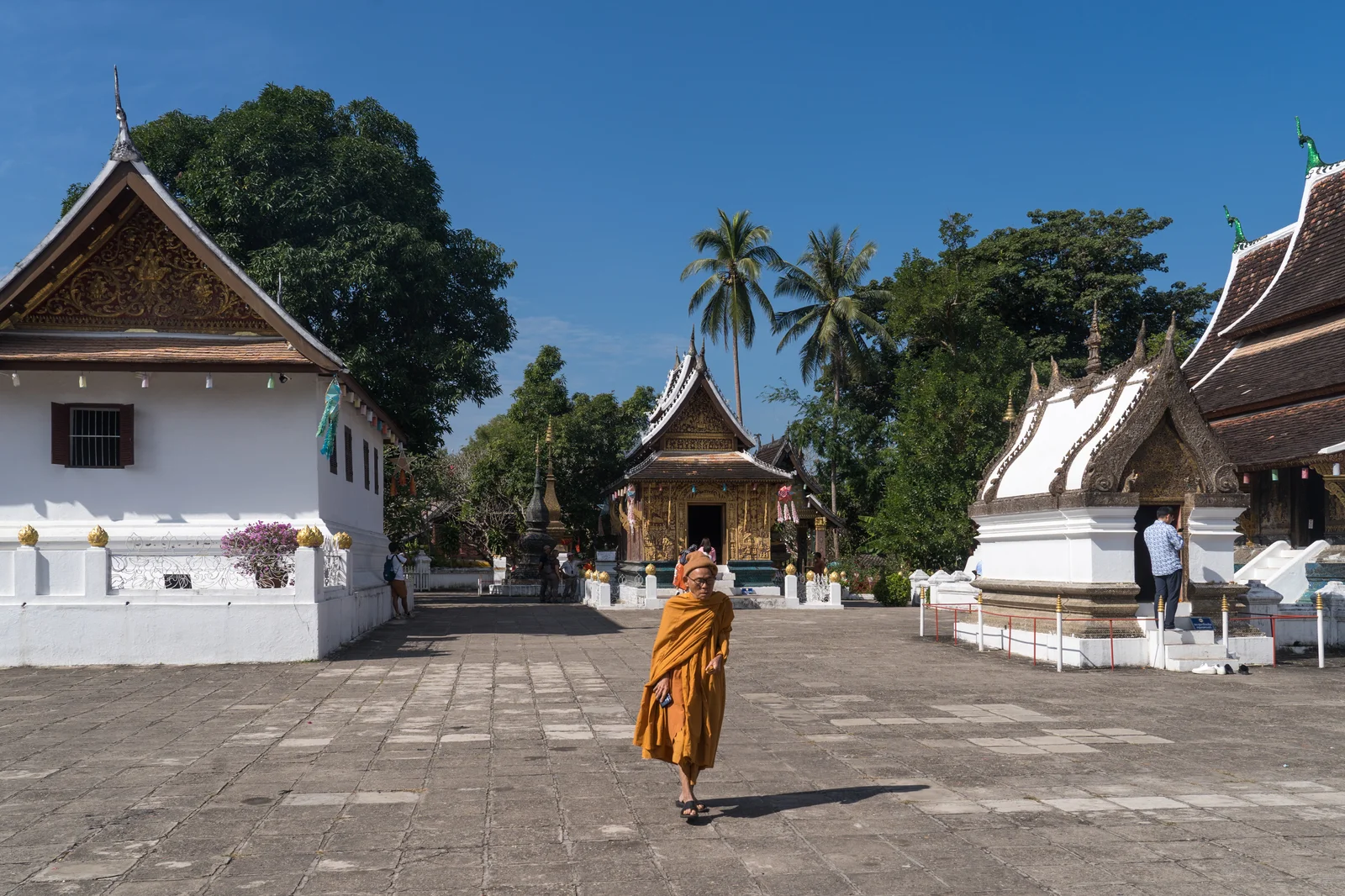 A saffron-robed Buddhist monk walking across the paved stone courtyard of Wat Xieng Thong temple in Luang Prabang, Laos, past whitewashed shrines and gilded roofs