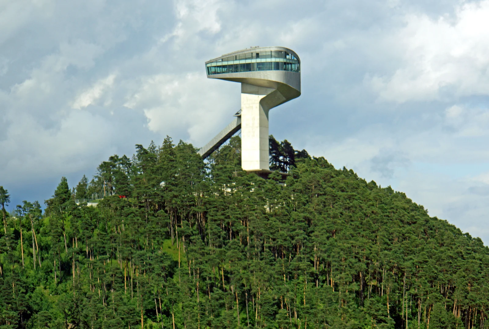 The Bergisel ski jump tower with its curved viewing-deck top rising above a densely forested green pine hillside under a partly cloudy sky