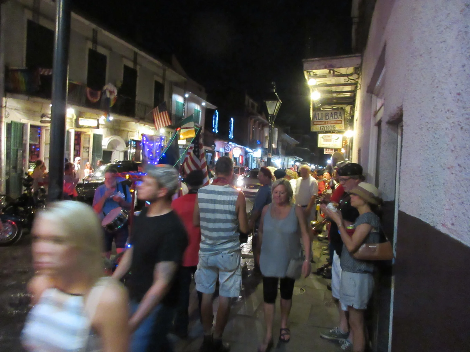 Crowds fill the Bourbon Street sidewalk at night, lit by neon bar signs, a gas-style street lamp, and rainbow flags draped from a balcony