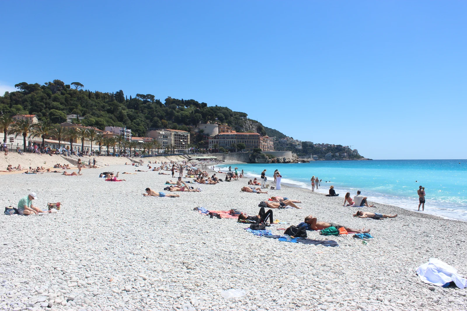 Sunbathers on the white pebble beach of the Promenade des Anglais in Nice, with turquoise sea and a wooded headland behind