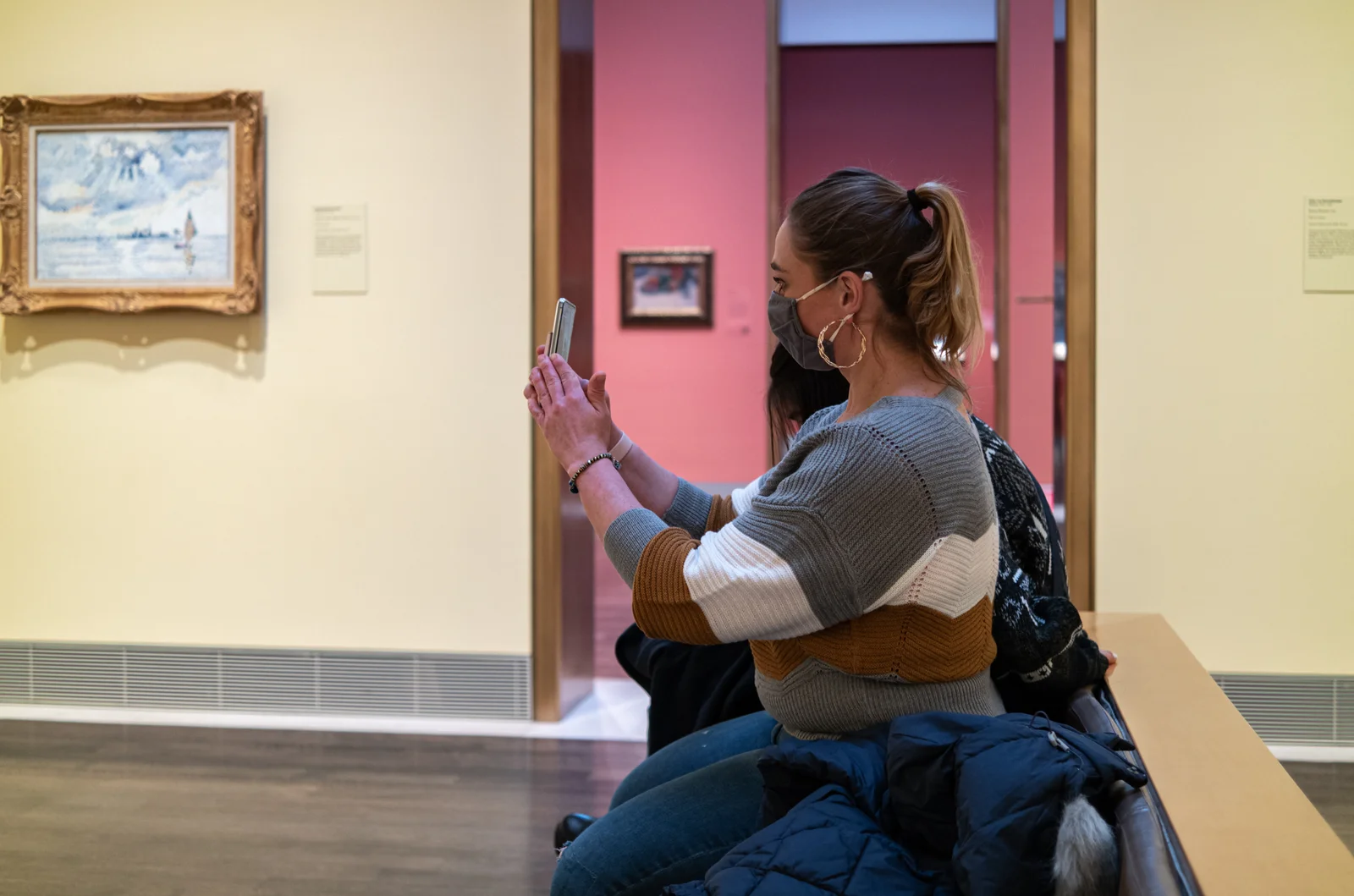 A seated museum visitor photographing a small framed painting on a gallery wall inside the Museum of Fine Arts, Houston