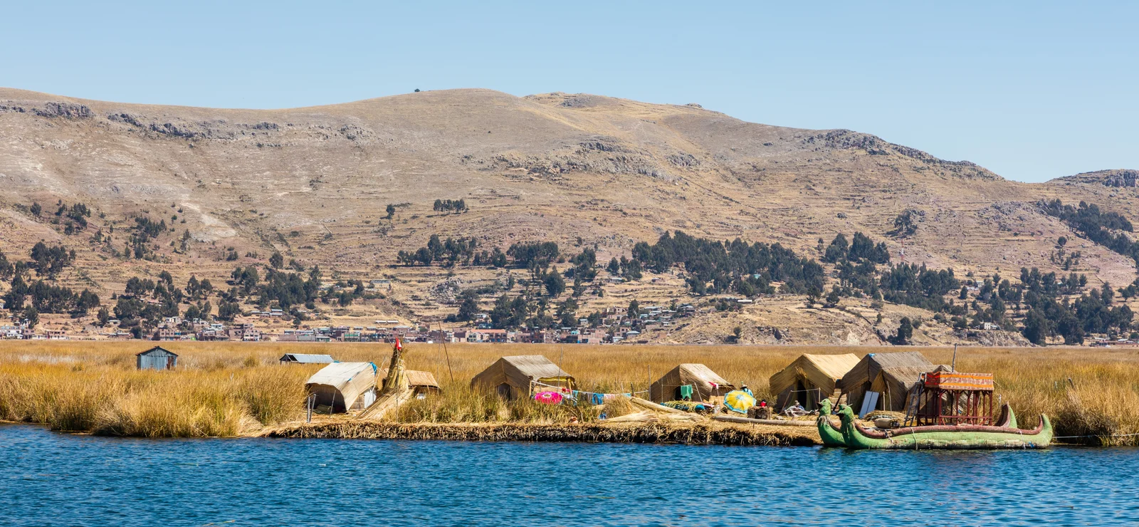 A floating island of dried golden reeds on Lake Titicaca holding thatched reed huts and a reed boat, with a bare tan hillside and a lakeside town across the water