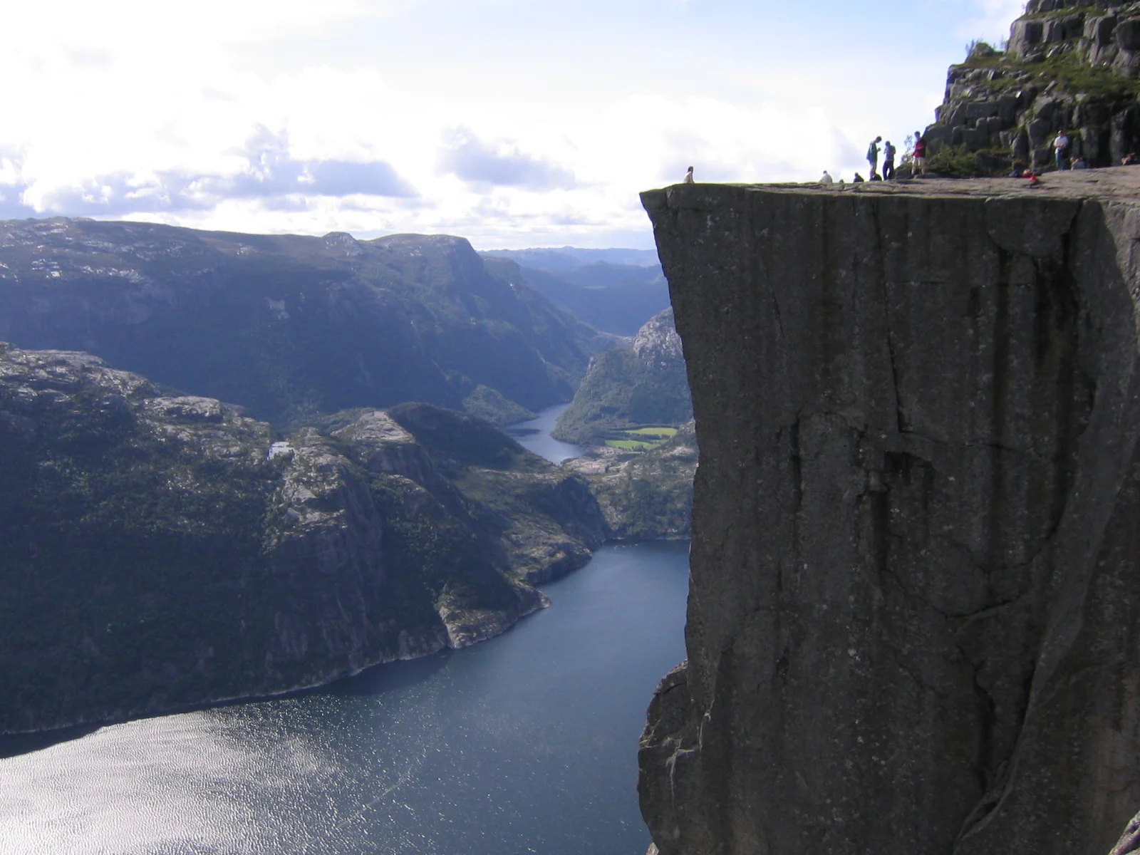 The Preikestolen cliff plateau seen from the side, with a few hikers standing near its edge high above Lysefjord