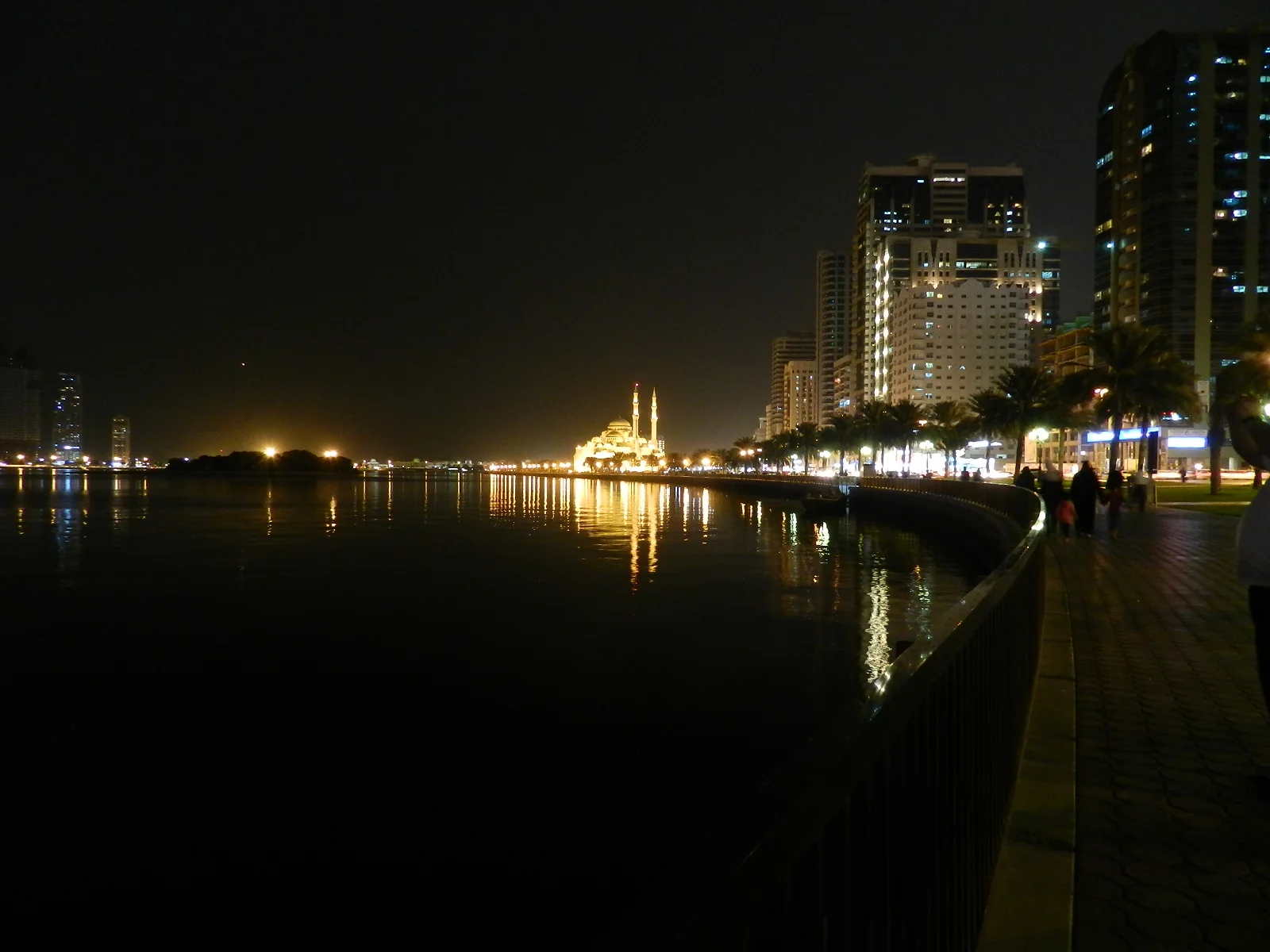 Al Noor Mosque floodlit gold at night across Khalid Lagoon, seen from the Buhaira Corniche with high-rise towers on the right