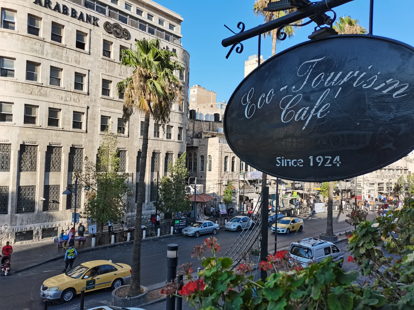 A downtown Amman street seen from a café terrace, with the grey-stone Arab Bank building, a fan palm, yellow taxis, and a hanging oval 'Eco-Tourism Café' sign