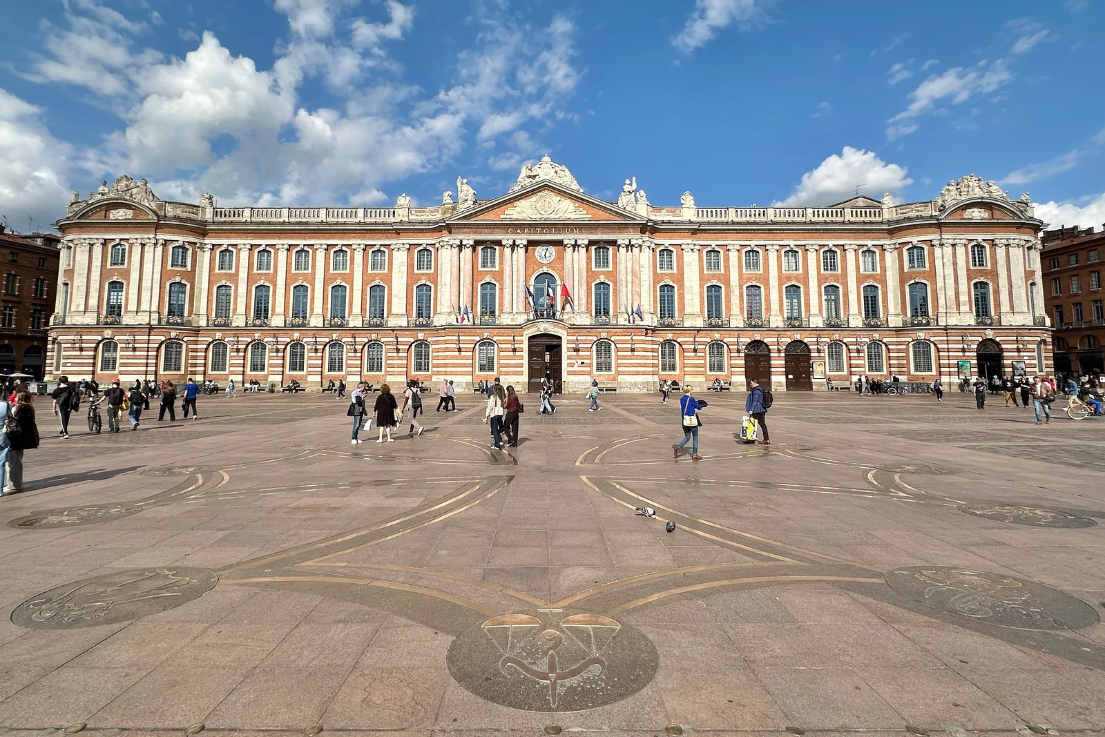The Capitole's long pink-brick facade seen head-on across the square, with the bronze Occitan cross inlaid in the paving in the foreground