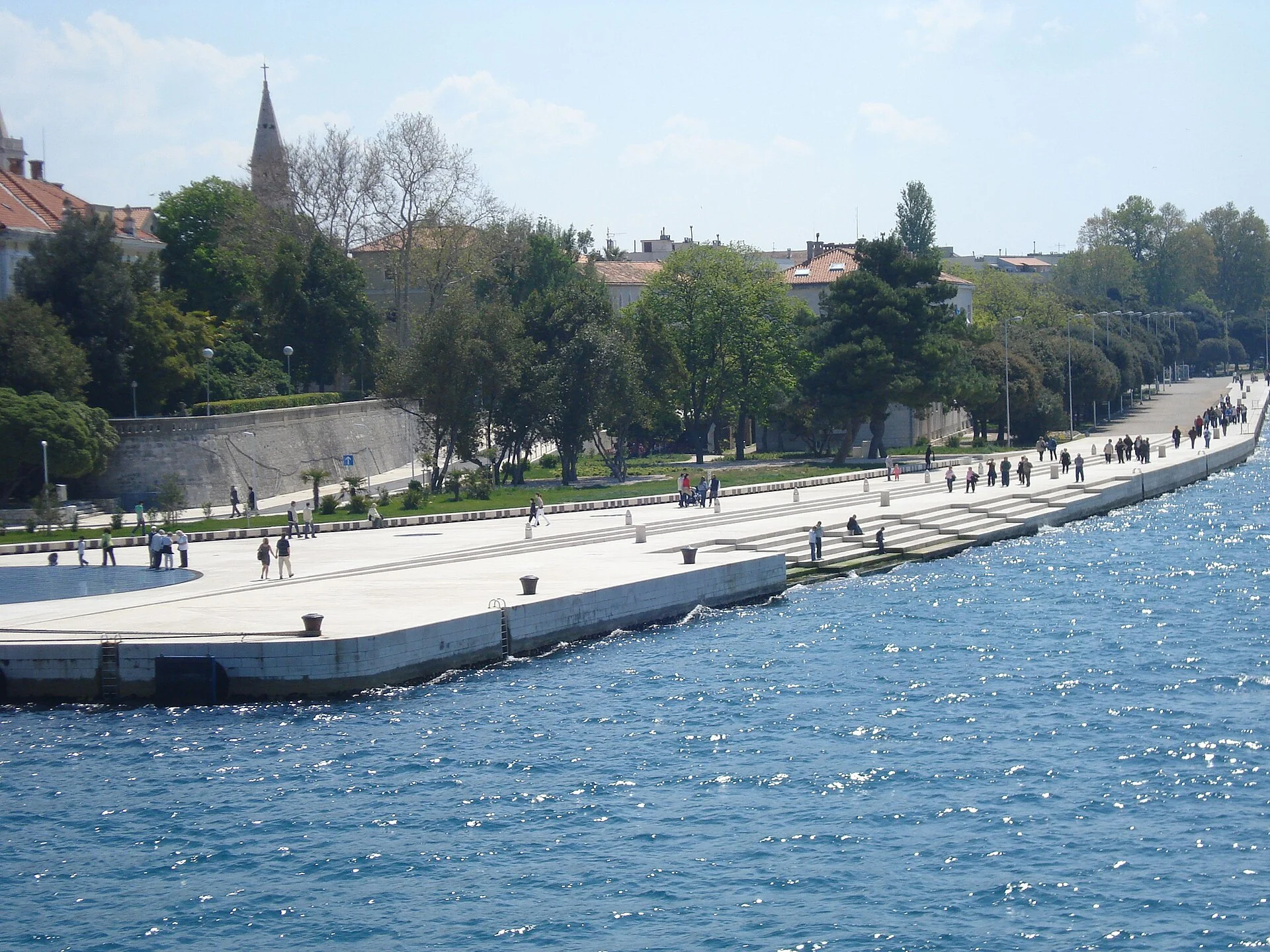 The white marble steps of Zadar's Sea Organ descending into the Adriatic beside the Riva promenade, with the bell tower of St. Anastasia rising over the old town