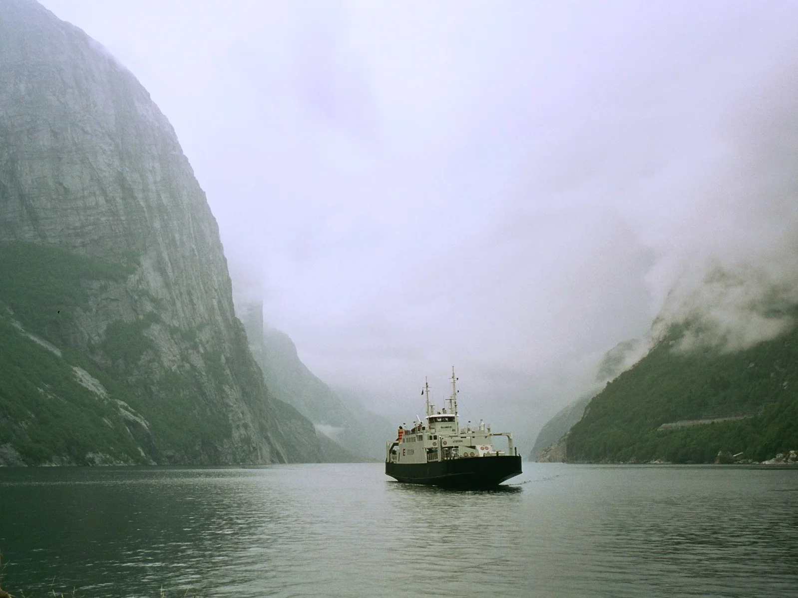 A small passenger ferry crossing the calm waters of Lysefjord between steep cliff walls on a misty morning