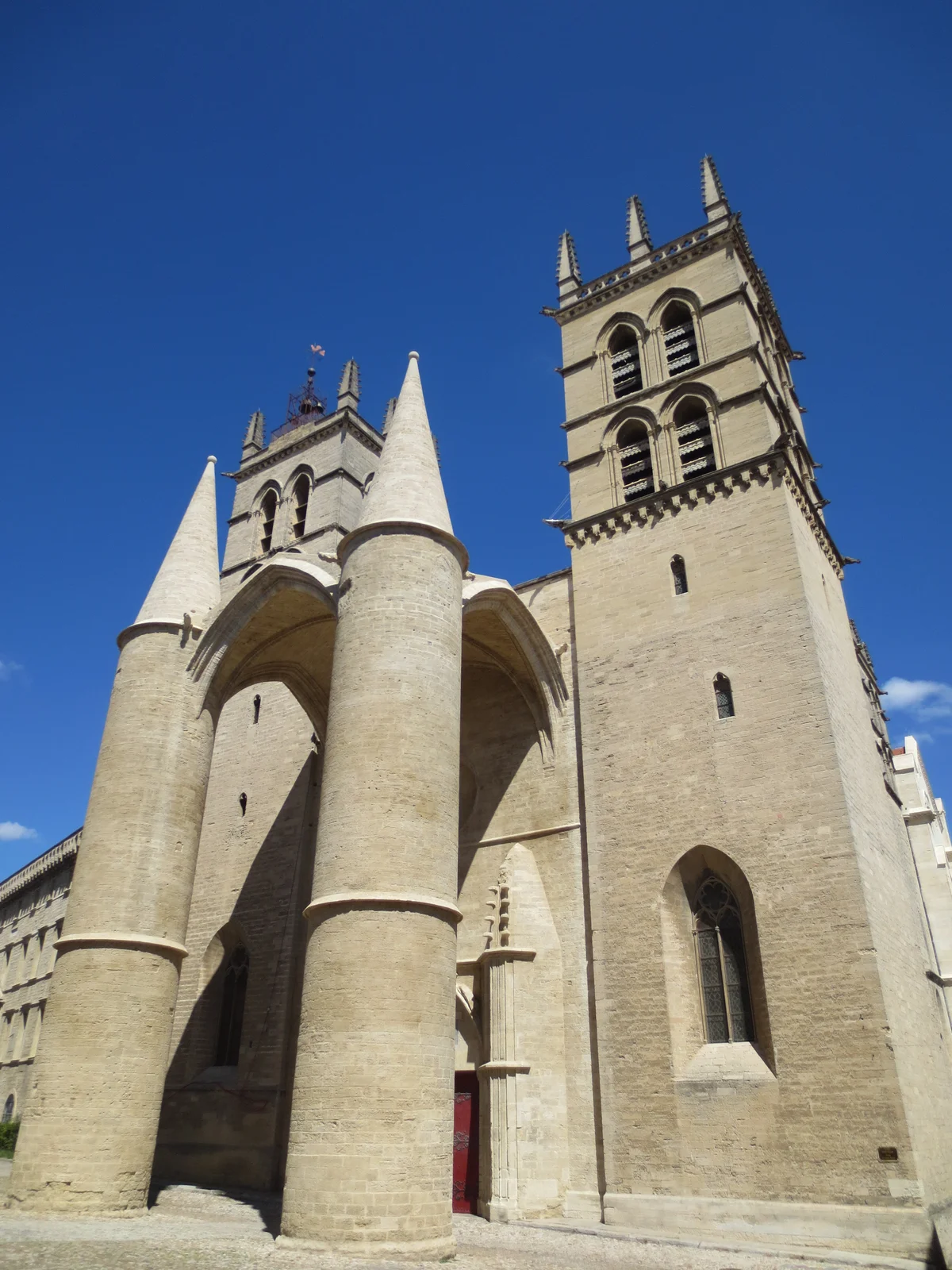 The west front of Montpellier's Saint-Pierre Cathedral, with its twin conical-canopied porch pillars and Gothic bell tower rising against a deep blue sky