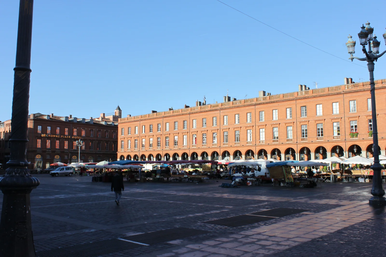 Covered market stalls with white awnings lining the red-brick arcaded houses on one side of Place du Capitole in low morning sun