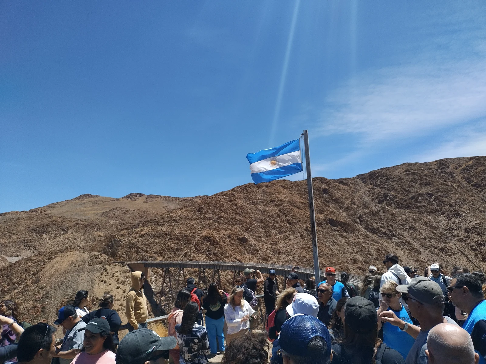 Tourists gathered beside an Argentine flag on a rocky ridge, with a steel railway viaduct curving across the barren brown hillside behind them