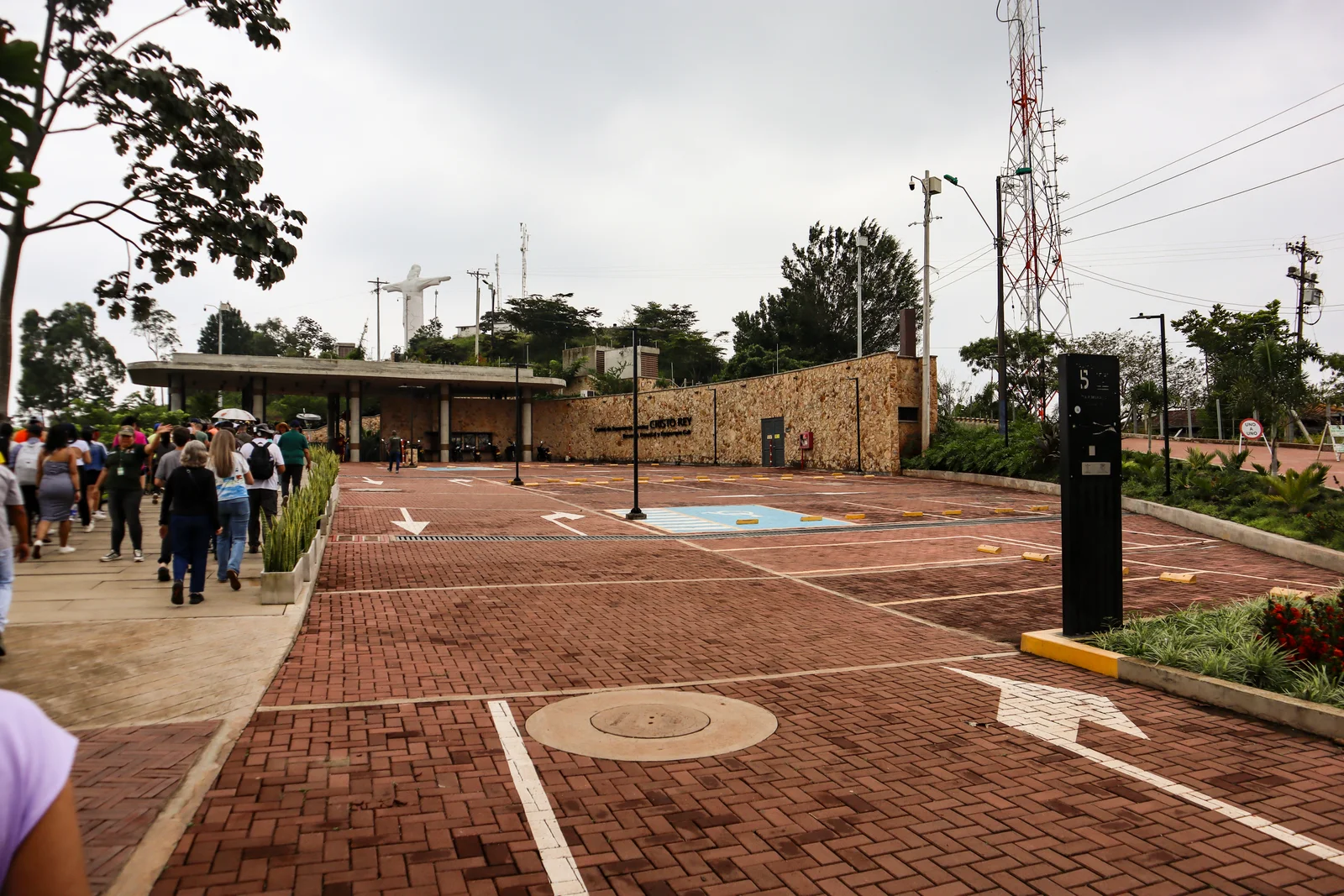 Wide brick-paved summit esplanade at the Cristo Rey ecopark, with visitors walking and the white statue small on the ridge beyond