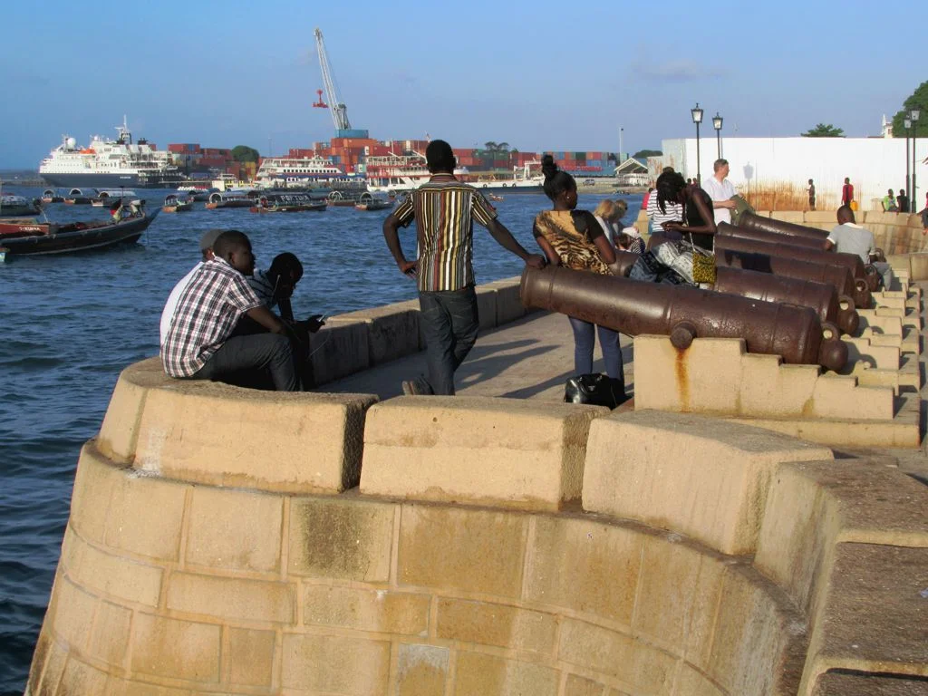 Row of old cannons along the Forodhani seafront wall in Stone Town, people sitting above the harbour