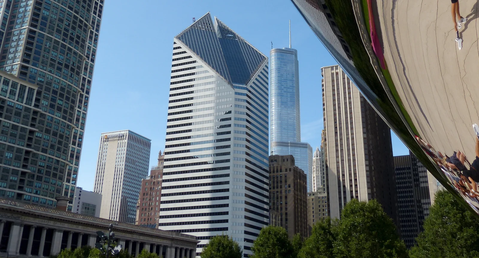 Downtown Chicago skyscrapers reflected beside the curved, mirror-polished flank of Cloud Gate, with visitors mirrored in the steel at right