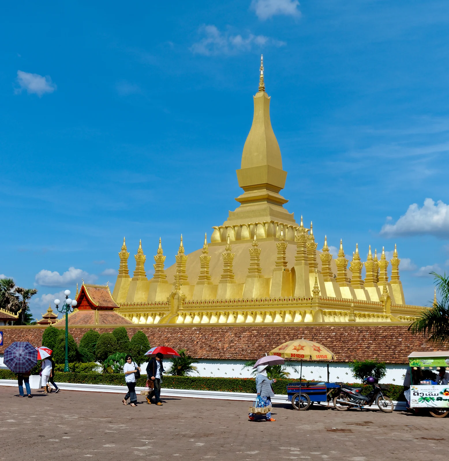 The golden stupa of Pha That Luang rising above its walled plaza in Vientiane, Laos, with people carrying parasols in the foreground under a blue sky