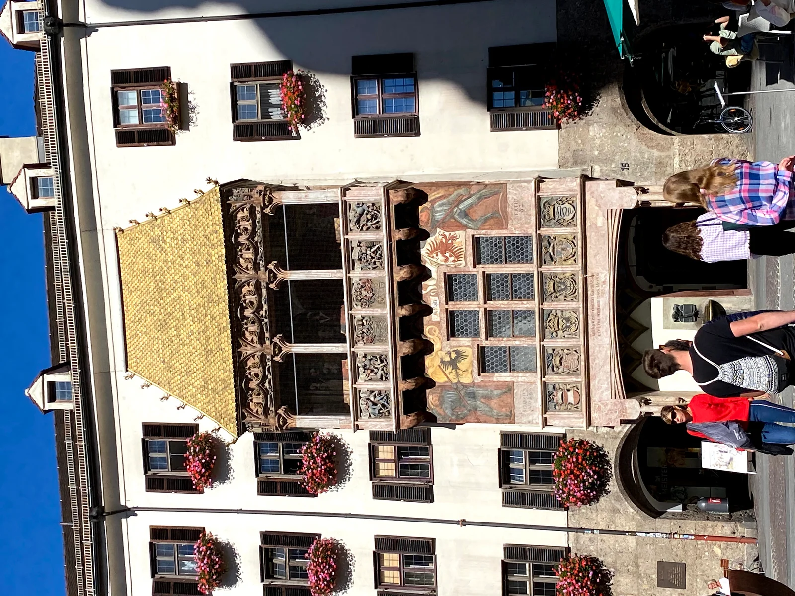 The Golden Roof's late-Gothic oriel balcony with its gilded shingled roof, carved stone parapet reliefs and painted heraldry, set into a cream townhouse facade with geranium window boxes