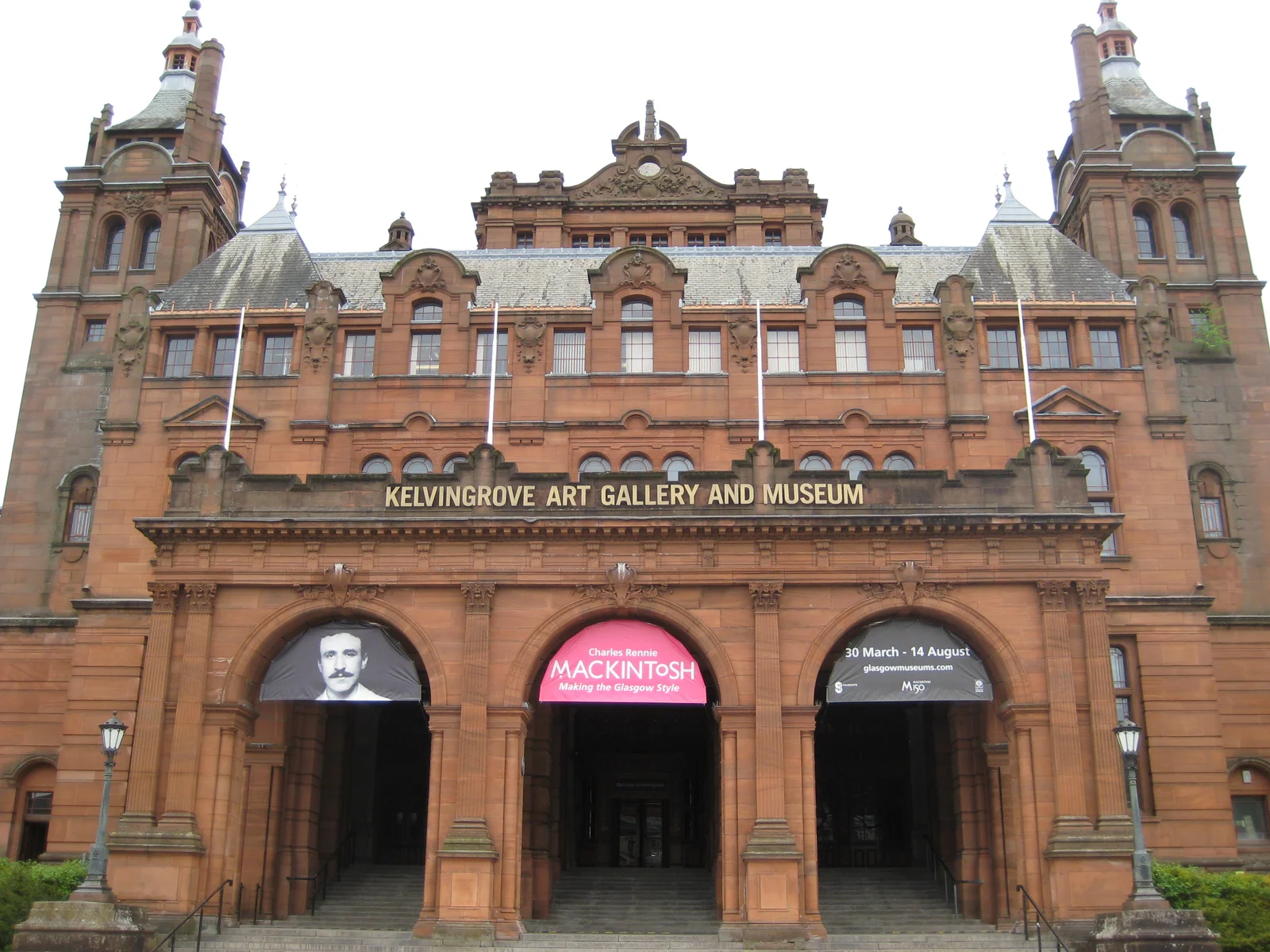 The red sandstone entrance facade of Kelvingrove Art Gallery and Museum, with twin towers and three arched doorways beneath an overcast sky