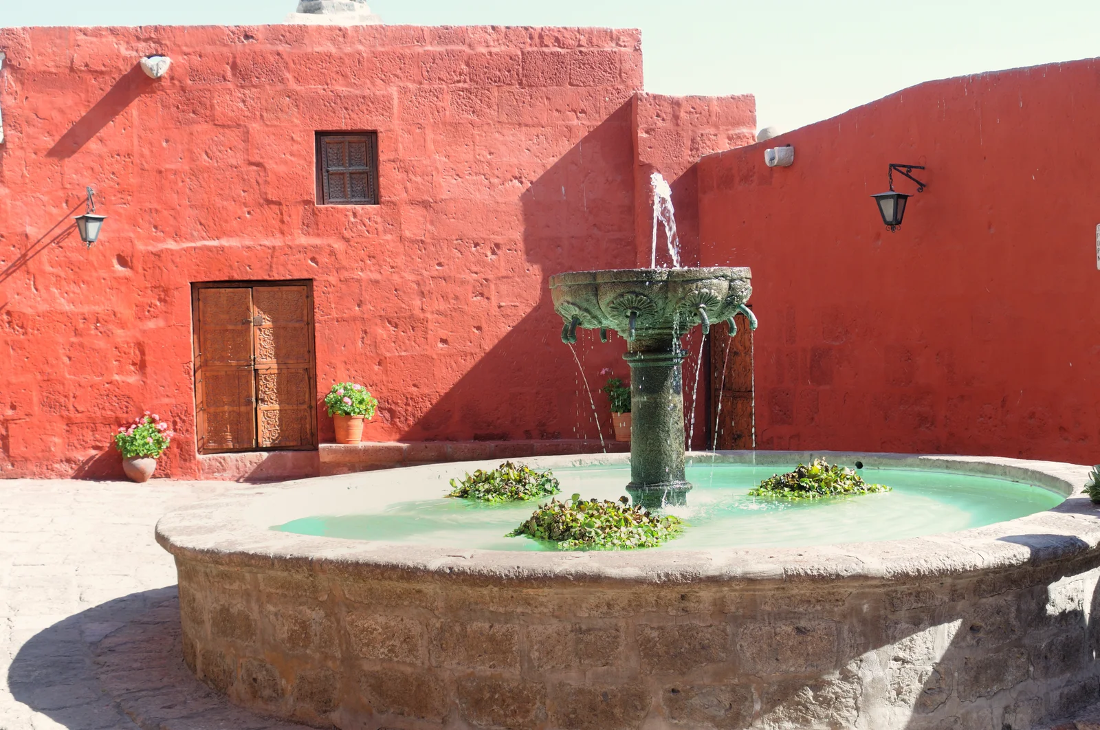 A weathered stone fountain running with water in a sunlit courtyard, surrounded by walls painted deep terracotta red with wooden shutters and potted geraniums
